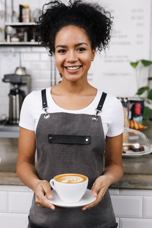 Young smiling waitress holding a cup of latte, standing at bar desk in coffeeshop