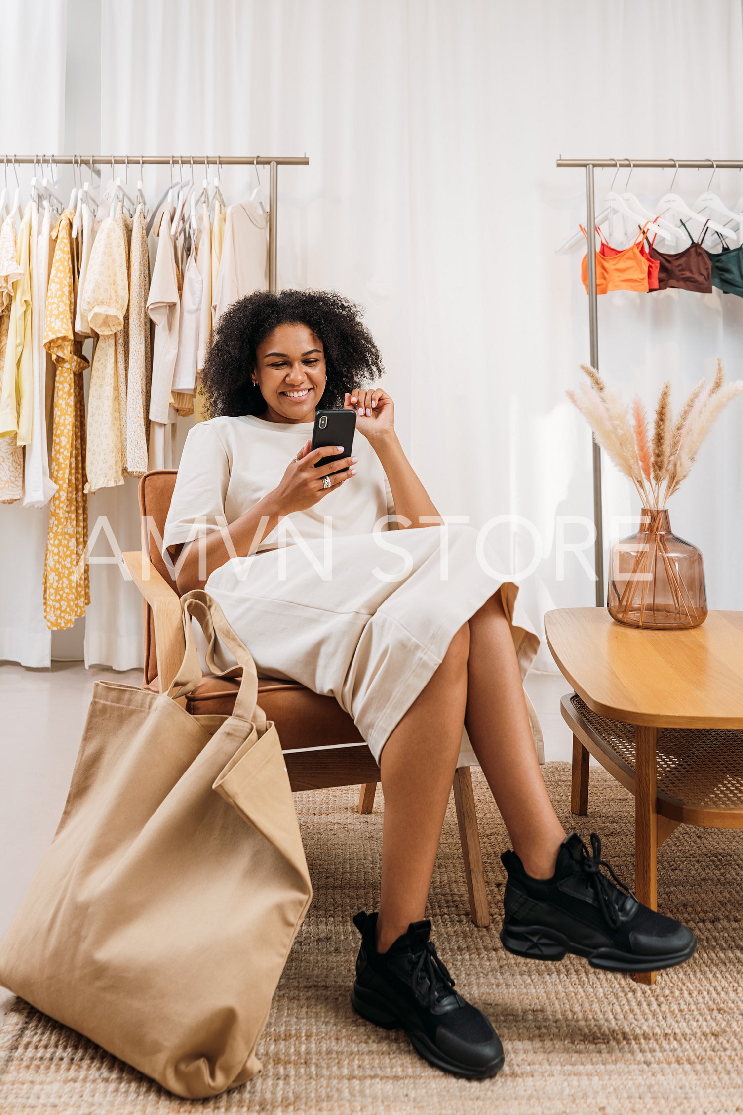 Smiling woman with mobile phone sitting on armchair in a clothing store. Customer relaxing in a boutique.