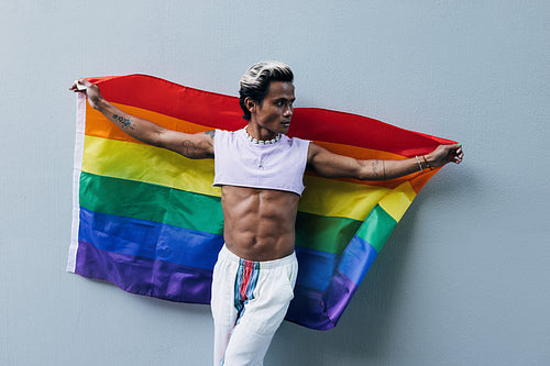 Muscular stylish guy holding an LGBT rainbow flag at a grey wall