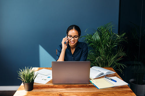 Smiling businesswoman sitting at a table talking on cell phone while working on laptop