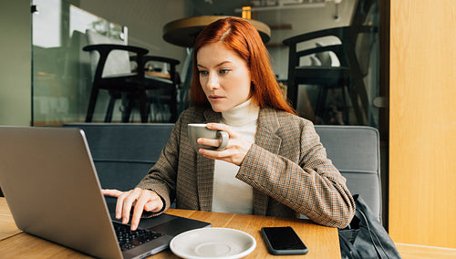 Woman working as a digital nomad in a cafe. Female entrepreneur with ginger hair holding a cup typing on a laptop