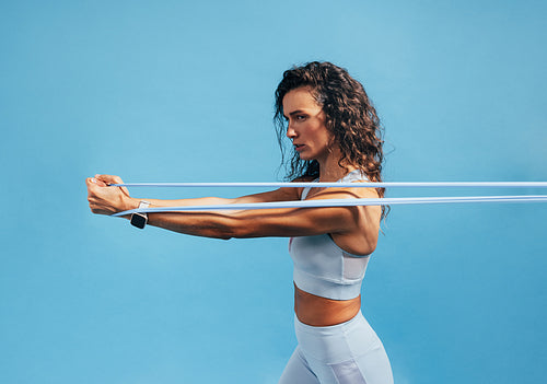 Side view of young fit woman doing intense training with resistance band on blue background