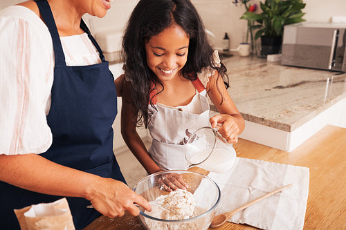 Smiling little girl and her grandma mixing flour in a bowl. Granddaughter pouring milk while granny whisking the flour.