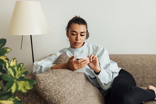 Young woman in casuals lying on a sofa in a living room holding a smartphone