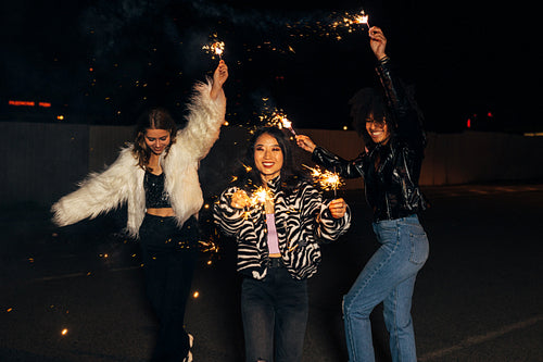 Three stylish girls having fun while walking with sparklers on city street at night