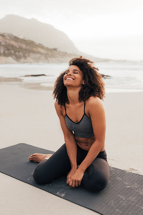 Happy woman in fitness wear sitting on a mat at the beach with closed eyes