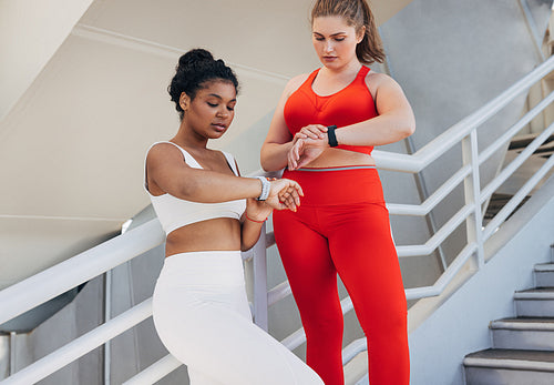 Two plus-size women in fitness attire are checking pulse during a workout. Young females in sports clothes are standing on stairs outdoors looking at smartwatches.