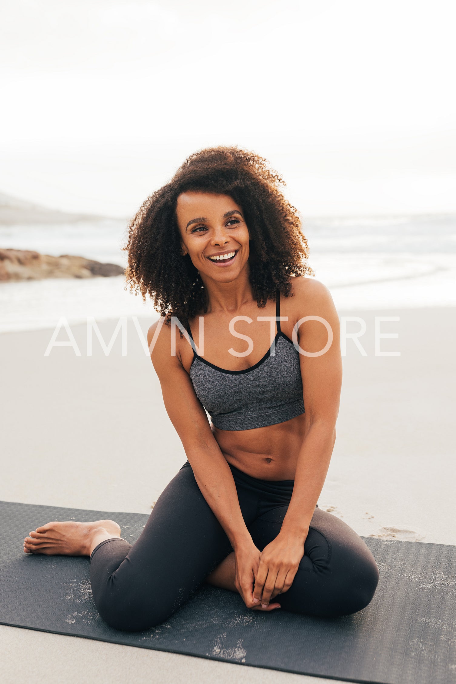 Happy woman with curly hair sitting on mat at beach. Smiling female relaxing after yoga exercises.