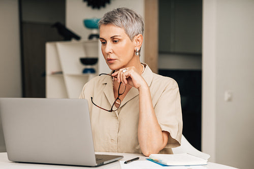 Mature woman in casuals sitting in the living room looking at laptop screen and holding spectacles