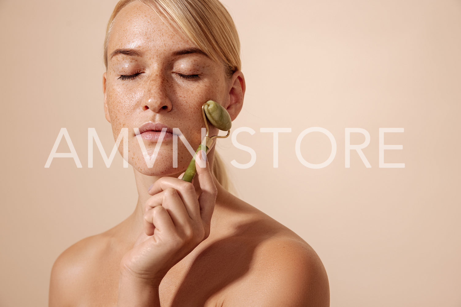 Blond woman with freckles using jade roller while standing with closed eyes against pastel background