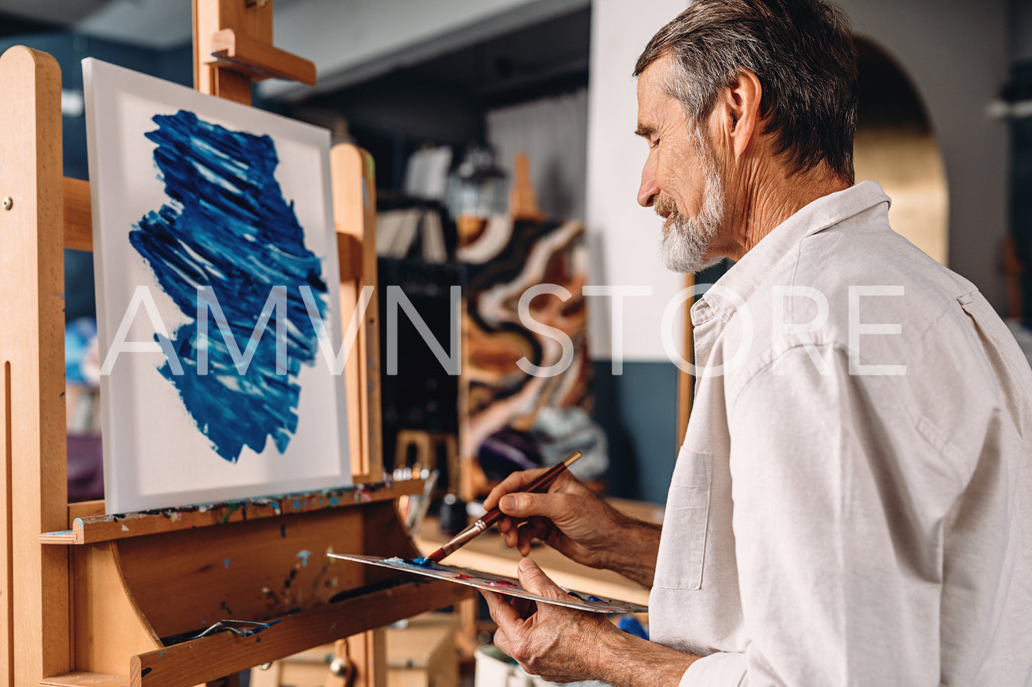 Concentrated bearded artist sitting in front of easel and mixing colors on palette with paintbrush	