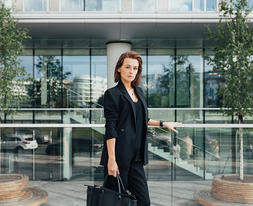 Serious and confident middle-aged woman wearing formal black clothes standing against a modern business building