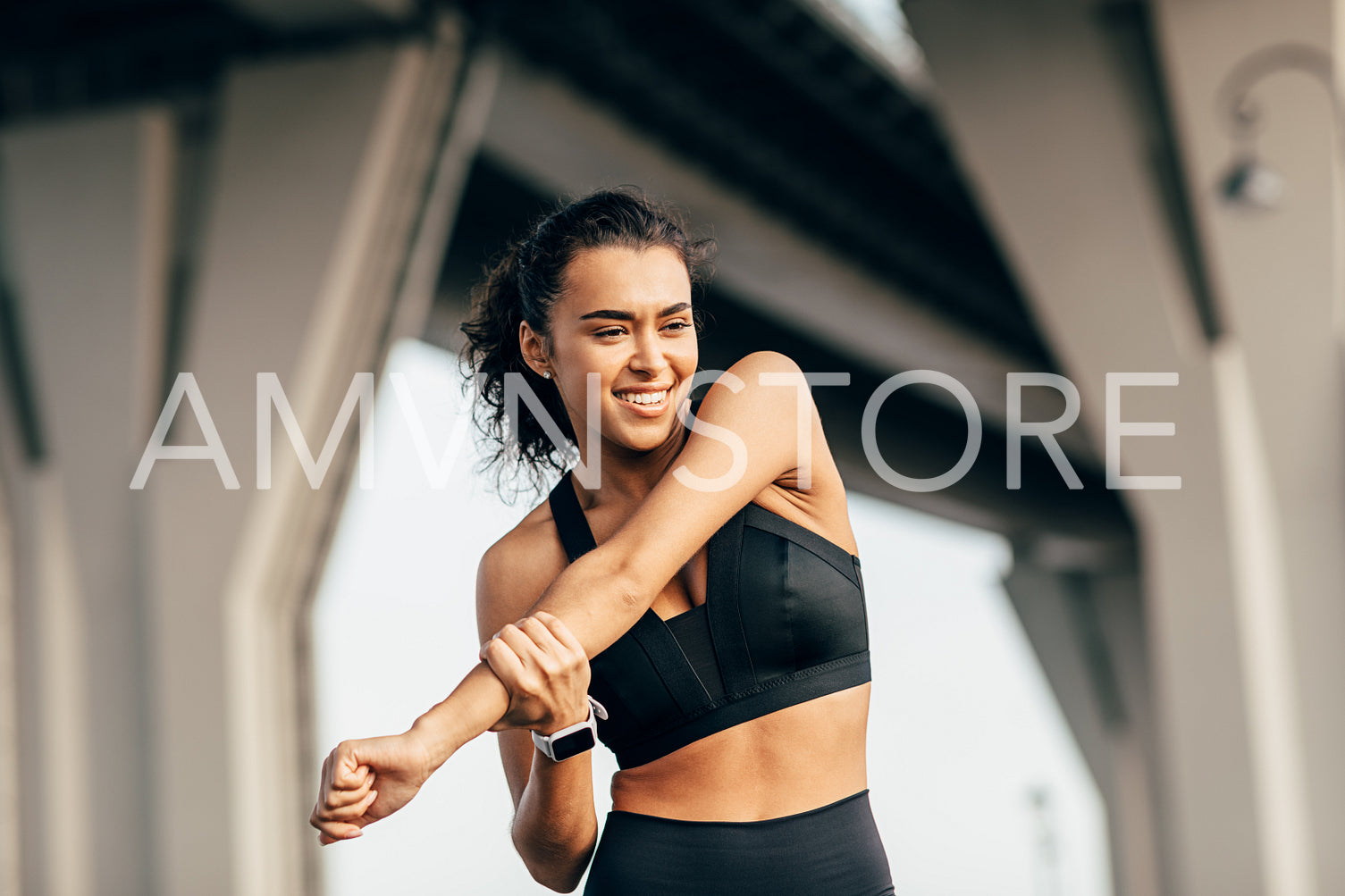 Woman athlete warming up her body before evening training under a highway. Smiling female stretching arm outdoors.	