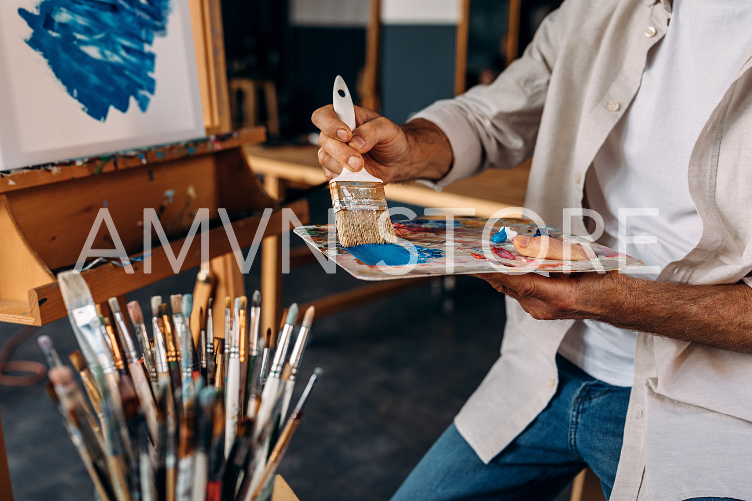 Cropped shot of artist sitting in front of an easel and dipping a large paintbrush into a colorful palette	