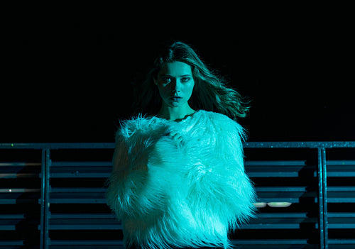 Woman in stylish fur coat standing at night under blue neon light