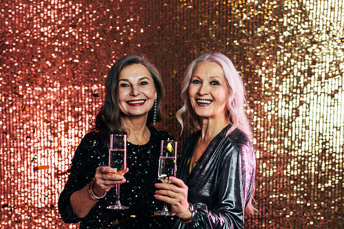 Stylish senior females standing together against glitter backdrop holding glasses of champagne