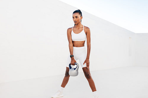 Slim woman practicing with a kettlebell in the white outdoor studio. Young female doing strength training outdoors.