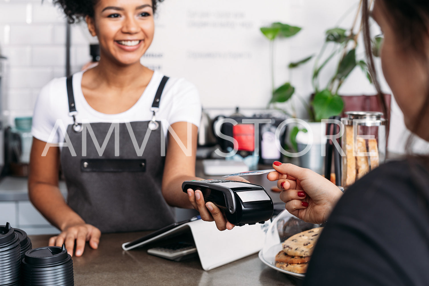 Smiling barista in apron holding POS terminal while customer paying by card