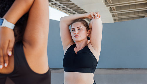 Portrait of a young woman stretching her hand with a fitness buddy. Two fitness females are exercising under bridge.