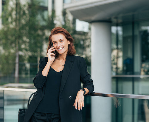 Cheerful businesswoman with ginger hair talking on a mobile phone. Smiling middle-aged female in formal clothes making a phone call outdoors.