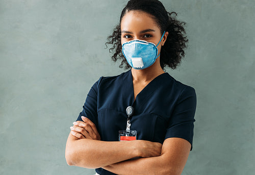 Young nurse in medical uniform with badge standing at wall with crossed arms