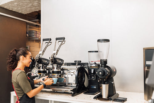 Female barista standing by coffee maker while working in cafe. Woman in apron preparing coffee.