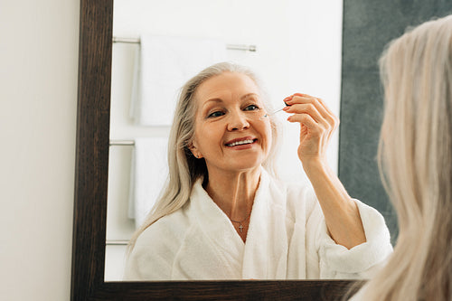 Smiling aged female applying the liquid facial treatment on face. Aged female with a hyaluronic dropper in bathroom.