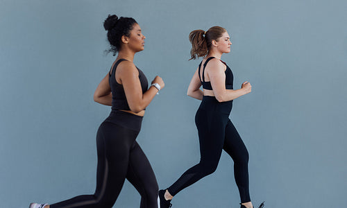 Side view of two plus size females running at grey wall. Two women in black fitness attire jogging outdoors.