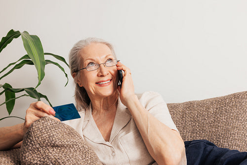 Smiling senior woman ordering through a cell phone. Aged female in eyeglasses holding a credit card while lying on a sofa.