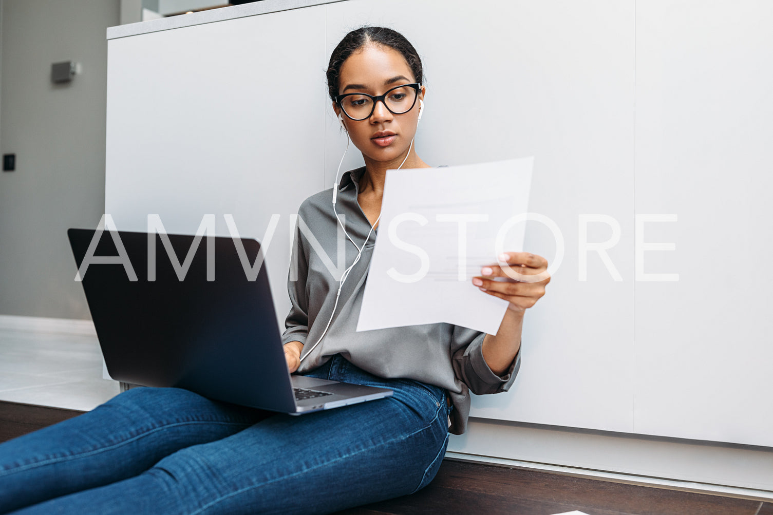 Young woman sitting on floor at kitchen counter with document in hand and using laptop	