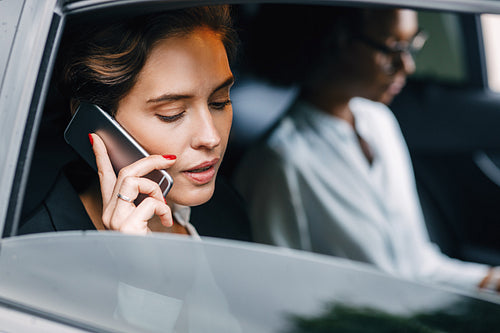 Businesswoman talking on a cell phone while sitting by a female colleague in a taxi