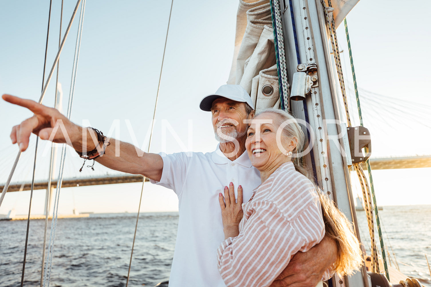 Mature man pointing into the distance and hugging his wife. Two smiling people enjoying a boat trip at sunset.	