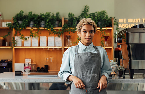 Confident female barista leaning on a counter looking at camera. Young woman in an apron working as a barista standing in a cafe.