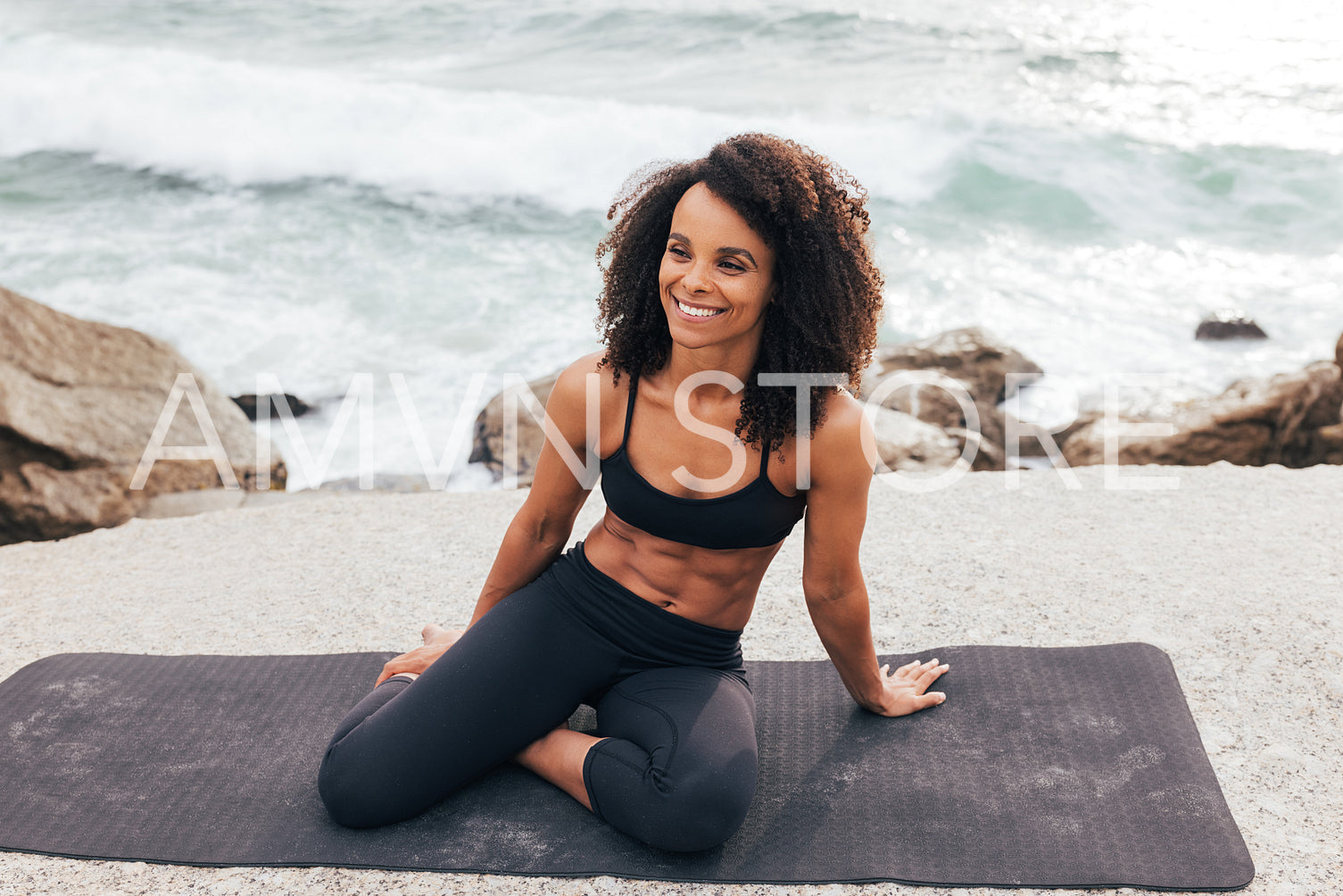 Happy muscular woman sitting on a yoga mat by ocean. Female with curly hair relaxing after outdoor training.