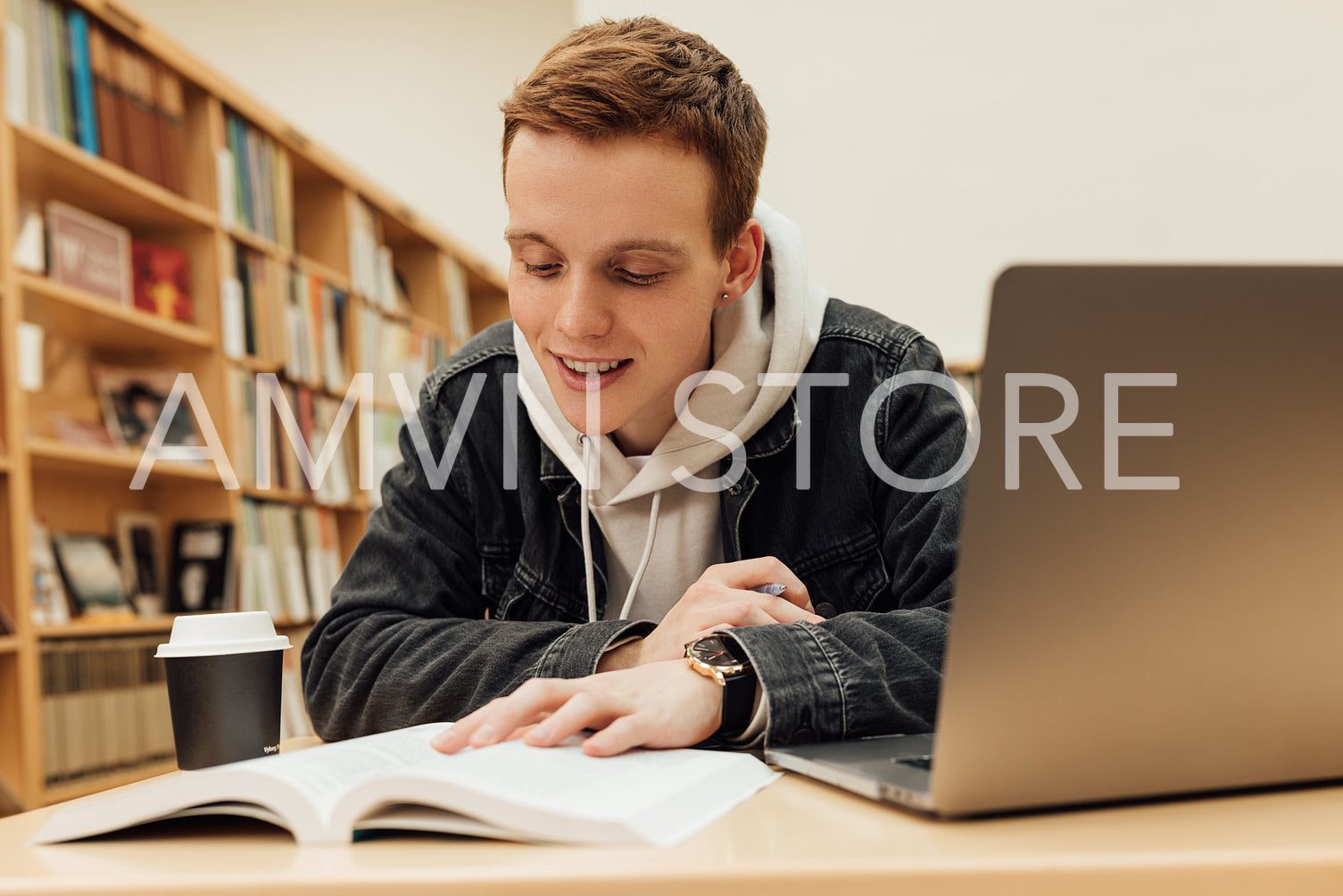 Smiling student with ginger hair preparing exams in library. Student reading from book while sitting at desk.