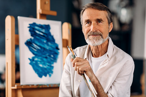 Portrait of bearded painter sitting against his picture and looking away