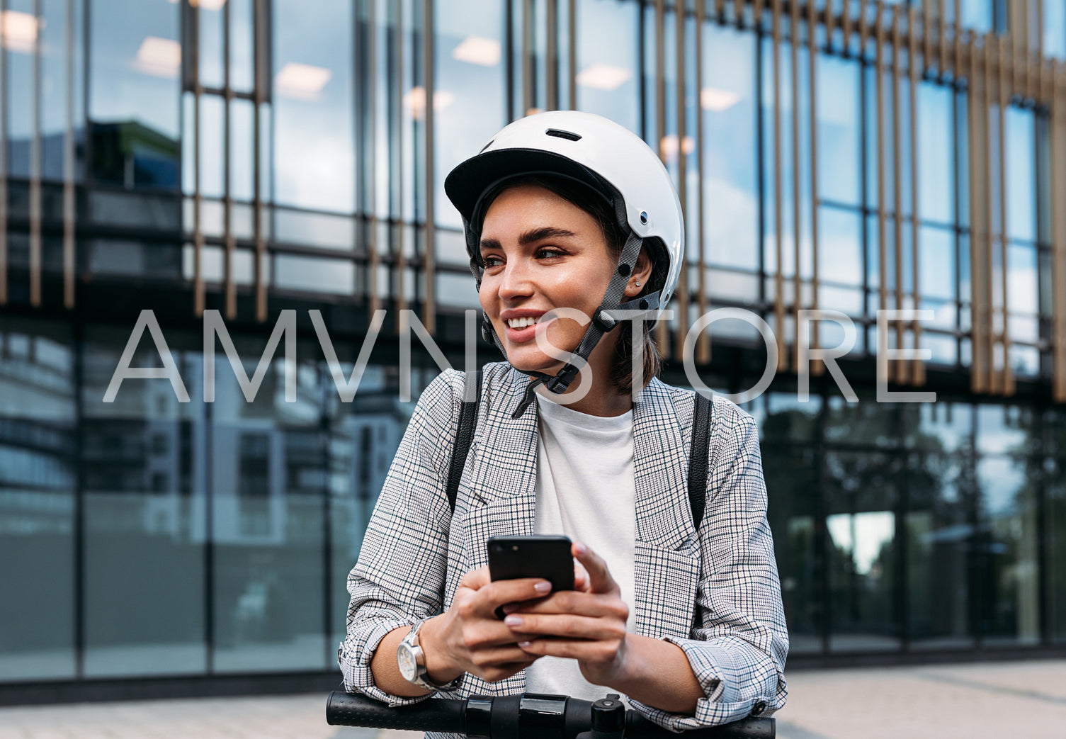 Smiling woman leaning on electric push scooter using a mobile phone while standing near an office building