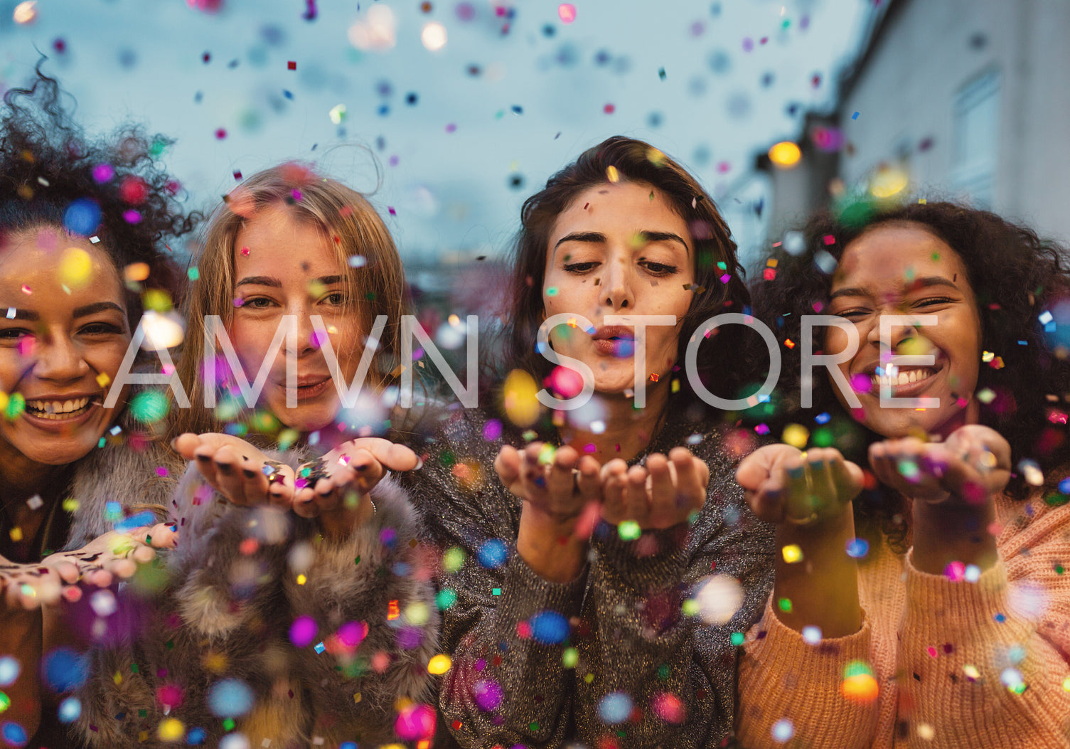 Young women blowing confetti from hands. Friends celebrating outdoors in evening at a terrace.	