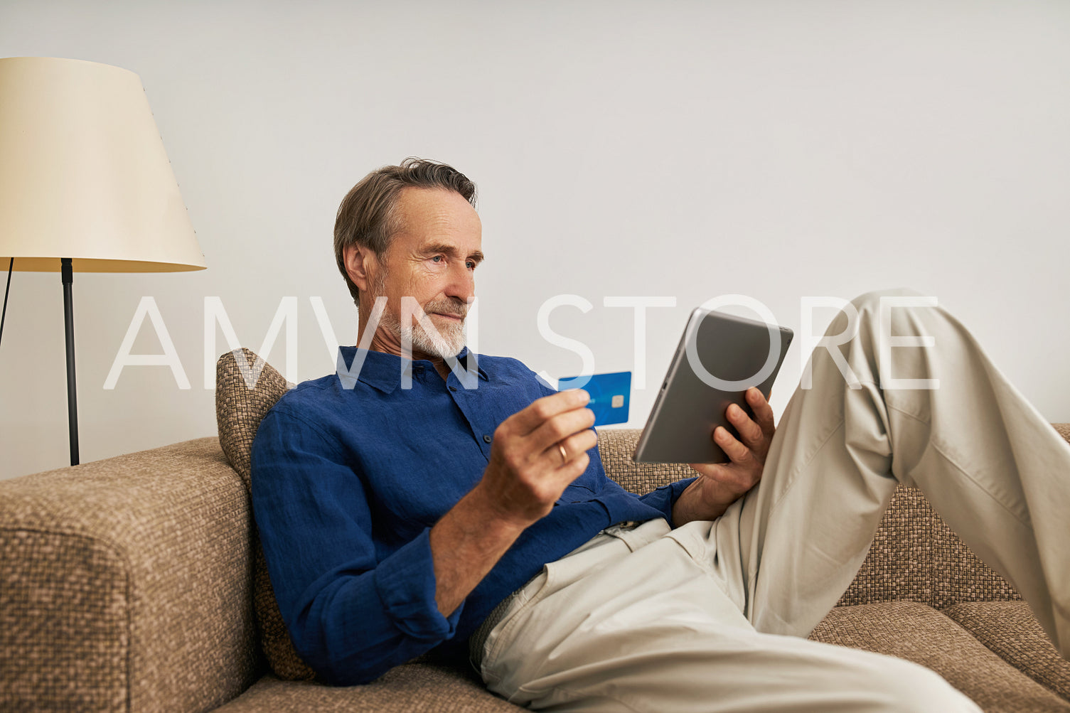 Handsome bearded man holding a digital tablet and credit card while sitting on a couch at home	