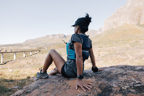 Side view of young woman relaxing on stone and looking on valley. Female in sports wear taking a break during trekking.