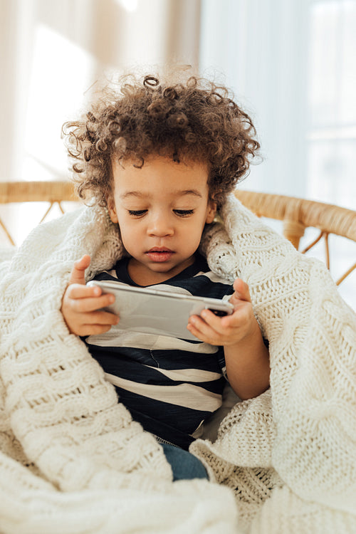 Little boy covered in blanket watching content on a smartphone at home