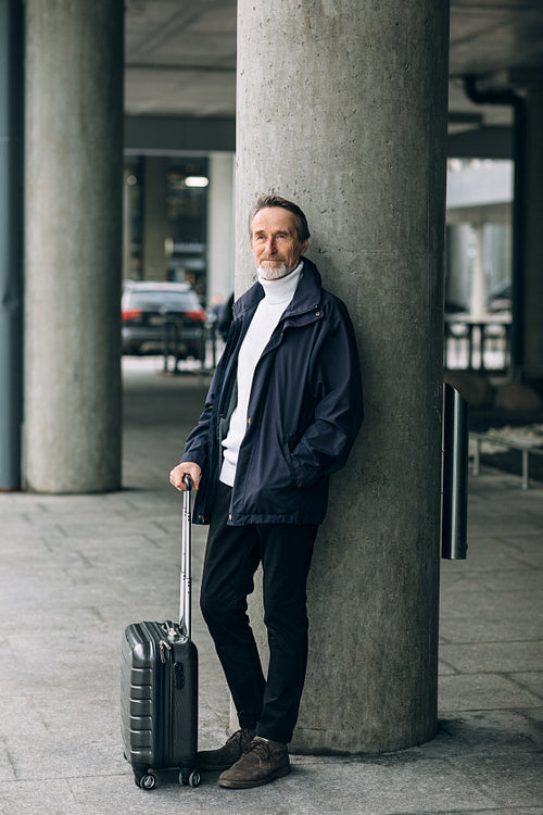 Senior man leaning on wall. Mature tourist standing outdoors with luggage.