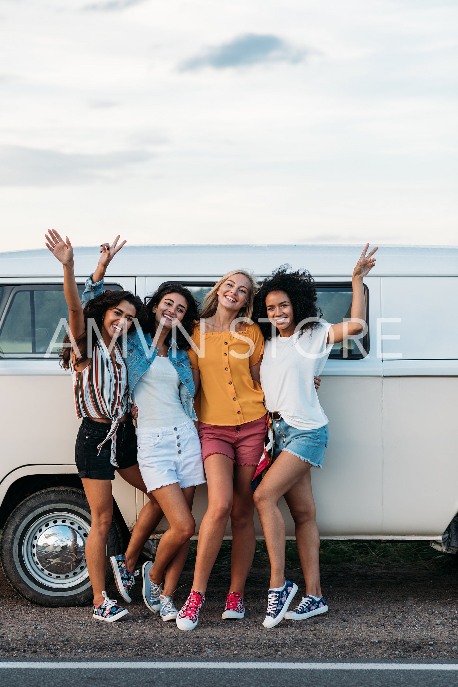 Four female friends in casuals standing at minivan on road raised hands up