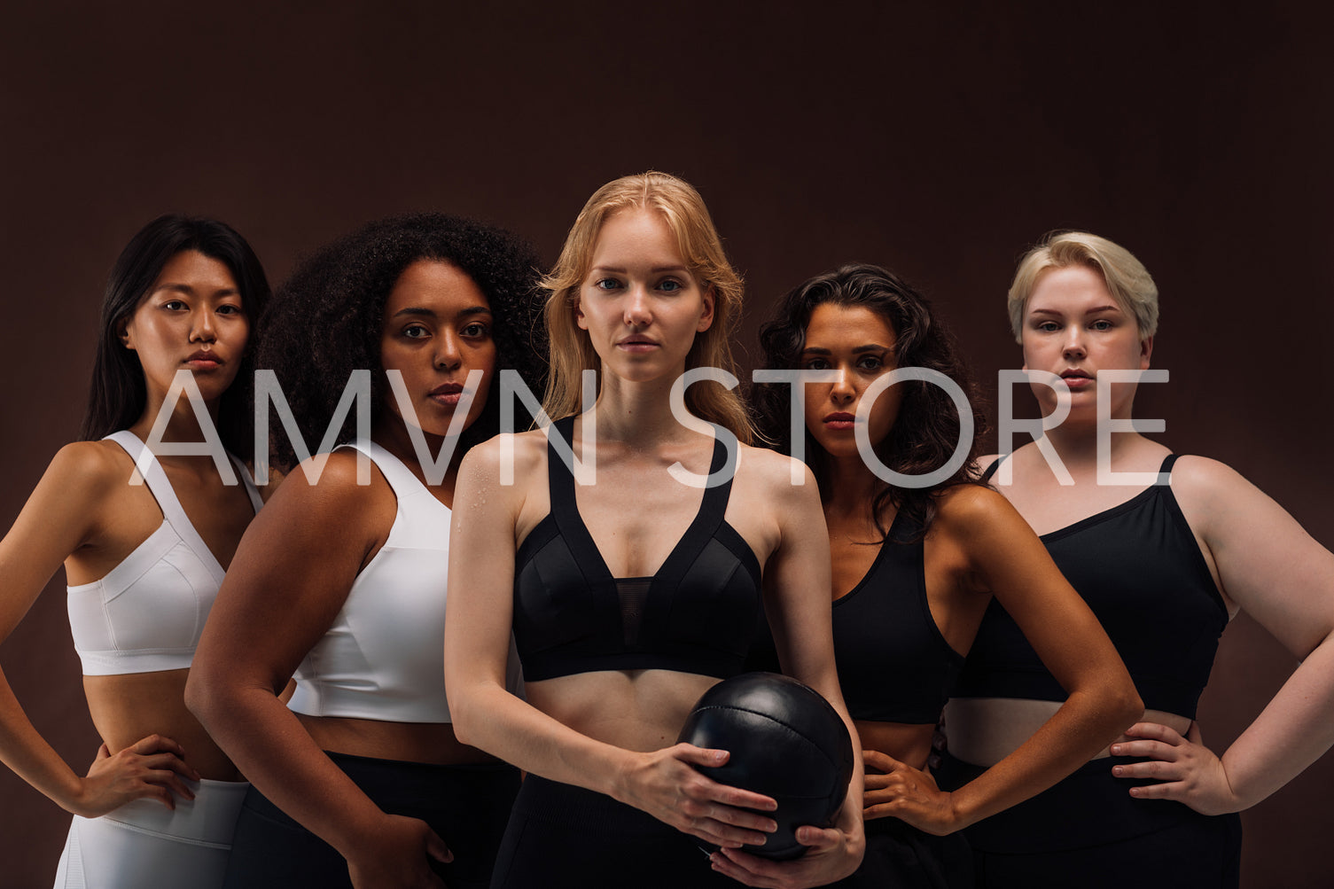 Five women in sports clothes standing together. Group of young females with different body type standing at brown background.