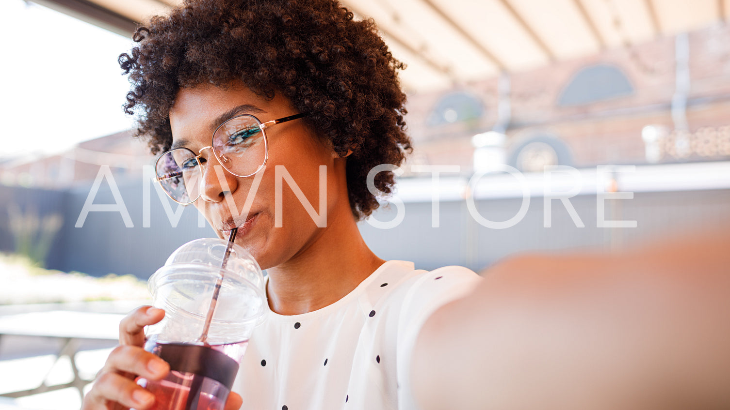 Close up of young woman drinking with straw while taking selfie on mobile phone, looking at frontal camera	