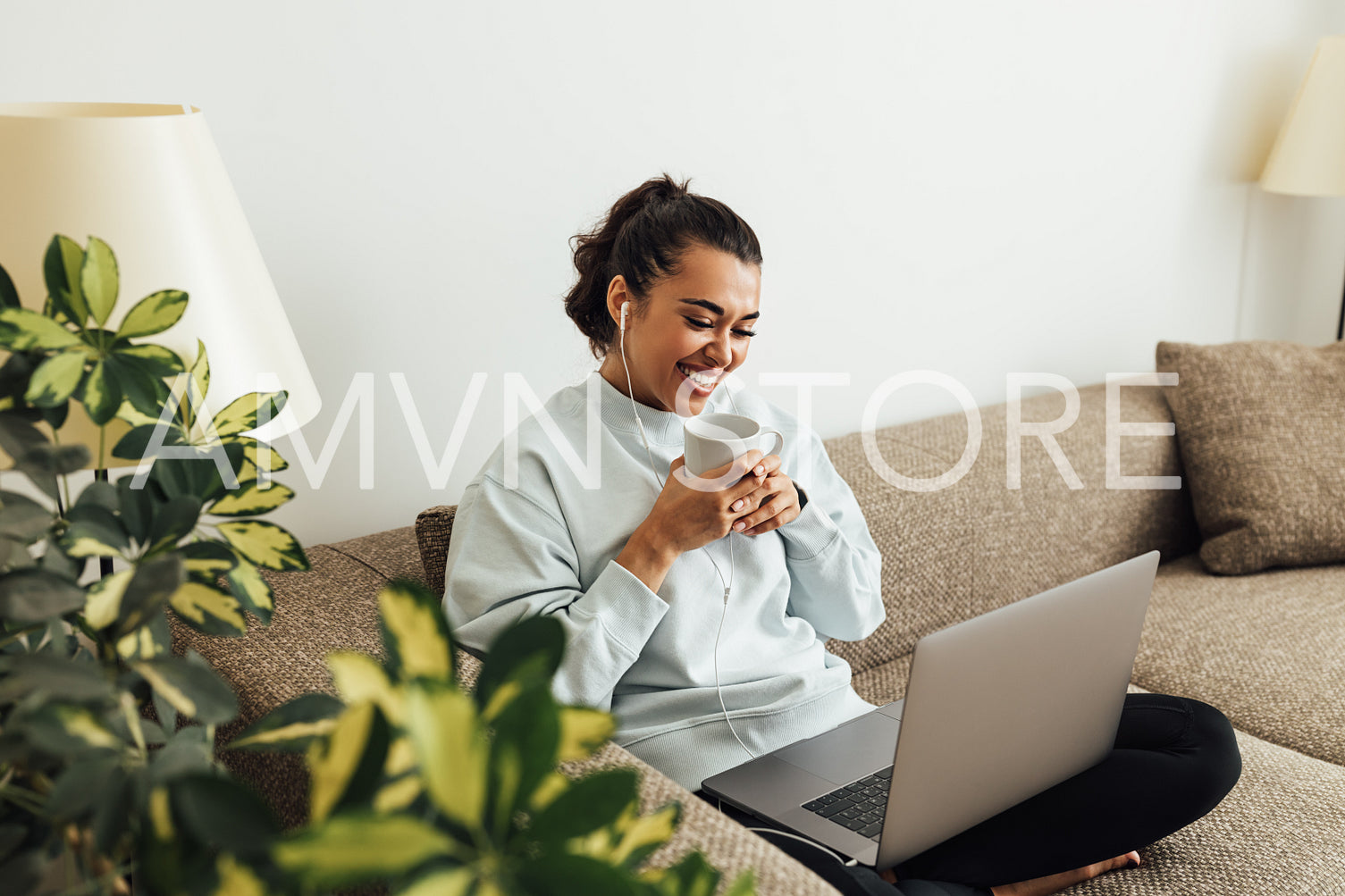 Young smiling woman holding a cup while sitting on a sofa with laptop on legs	