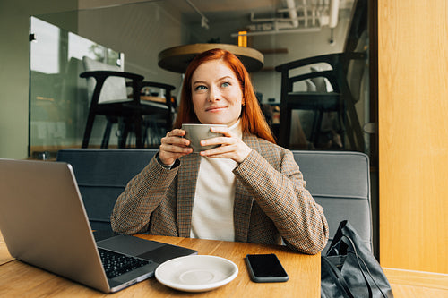 Smiling redhead woman in formal wear drinking coffee looking at window in cafe