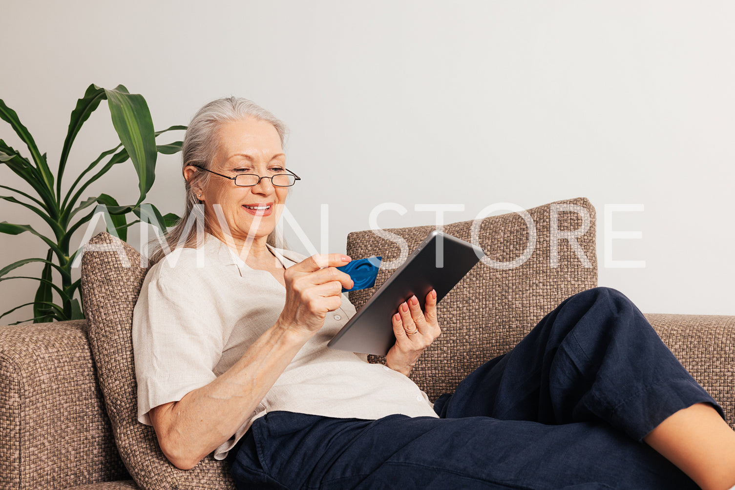 Smiling aged woman with a credit card. Mature female in eyeglasses holding a digital tablet while lying on a sofa at home.