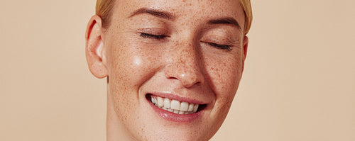 Close up highly detailed studio shot of a young female with freckles. Portrait of a smiling woman with blond hair and smooth skin against a beige background.