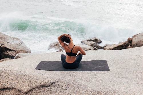 Rear view of young woman flexing her neck while sitting on yoga mat by ocean